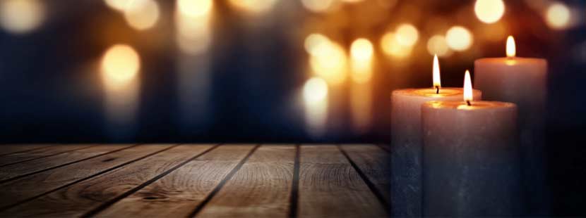 Drei brennende Kerzen auf einem Holztisch vor dunklem Hintergrund, in dem noch mehr Lichter leuchten. Trauerfall Checkliste Österreich.