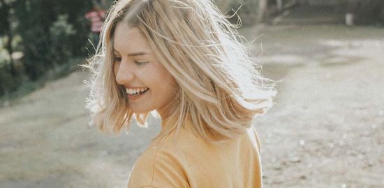 Frisurentrends 2023: Eine junge Frau mit blondem, schulterlangem Haar steht in einem sonnigen Garten und lächelt. Sie trägt ein gelbes Shirt.