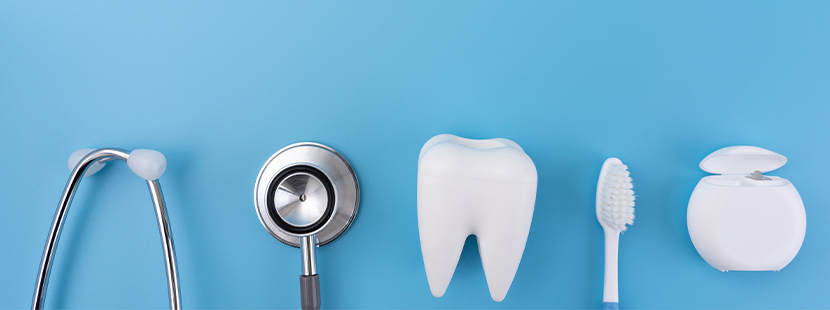 Ein Stethoskop, ein Zahn aus Plastik, eine Zahnbürste und eine Packung Zahnseide liegen auf einem blauen Hintergrund. 